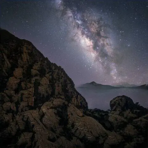 Vista nocturna de la Vía Láctea sobre un paisaje montañoso, con rocas en primer plano y un cielo estrellado que muestra colores vibrantes y detalles de la galaxia. Ideal para amantes de la astronomía y la fotografía de paisajes.