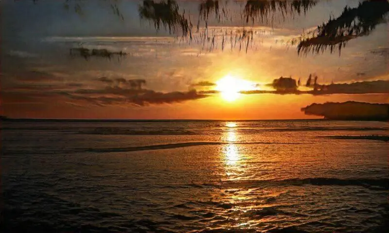 Atardecer sobre el mar con tonalidades naranjas y doradas, reflejándose en las aguas tranquilas, rodeado de nubes y vegetación. Ideal para ilustrar la belleza natural y los paisajes costeros.