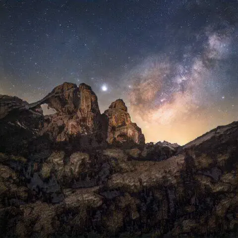 Paisaje nocturno de montañas con la Vía Láctea iluminando el cielo estrellado, destacando formaciones rocosas en primer plano y un ambiente sereno.