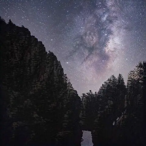 Vista nocturna de un cañón rodeado de árboles, con la Vía Láctea brillando en el cielo estrellado. La imagen muestra formaciones rocosas imponentes y un reflejo en el agua del cañón. Ideal para amantes de la naturaleza y la astronomía.