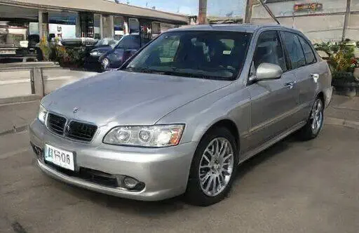 Automóvil sedan plateado con llantas deportivas y diseño moderno, estacionado en un área urbana con plantas y otros vehículos de fondo. Ideal para quienes buscan un coche con estilo y rendimiento.