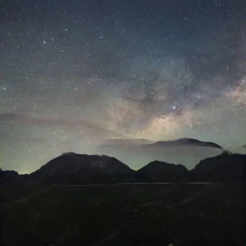 Panorama de un cielo estrellado con la Vía Láctea visible sobre montañas en un paisaje natural. La imagen muestra una mezcla de estrellas brillantes y nebulosas, creando un ambiente sereno y majestuoso.
